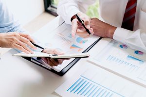 Close-up of two business professionals discussing financial and analytical charts. One person is holding a pen and a notebook, while the other is pointing at graphs and charts on a clipboard and table. The image highlights collaboration and the use of integrated HR and payroll systems to enhance workforce analytics.