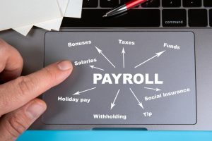 Close-up of a laptop touchpad with the word 'PAYROLL' in the center, surrounded by arrows pointing to terms like 'Bonuses,' 'Salaries,' 'Taxes,' 'Funds,' 'Social insurance,' 'Tip,' 'Withholding,' and 'Holiday pay.' A person's finger is pointing at the touchpad, symbolizing the various components managed by integrated HR and payroll systems.