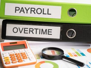 Binders labelled payroll and overtime on a desk with charts, calculator for small business payroll management.